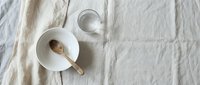 Minimalist white ceramic bowl with wooden spoon and glass of water on textured linen fabric background for kitchen styling and food photography.