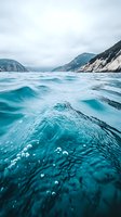 Turquoise ocean waves with air bubbles beneath surface against mountainous coastline background under cloudy sky, dramatic seascape perspective creates depth and movement.