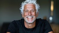 Mature Caucasian man with white hair and beard wearing black t-shirt, showing genuine happy smile against dark background. Natural lifestyle portrait for aging and wellness concepts.