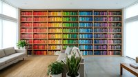 Modern home library with rainbow-colored book spines arranged by color creating vibrant wall display in contemporary living room with natural light.