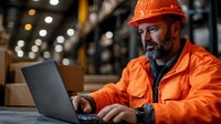 Warehouse supervisor in orange safety workwear and hardhat working on laptop computer while managing logistics and inventory in industrial storage facility.