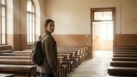 Young woman with backpack standing in empty vintage classroom with wooden benches and large windows creating warm natural lighting atmosphere.