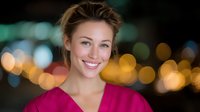 Young Caucasian female healthcare worker in pink medical scrubs smiling warmly against blurred city lights background, representing wellness and professional care.