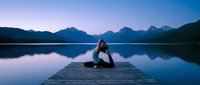 Young woman practicing yoga meditation pose on wooden dock at serene mountain lake during peaceful twilight hour for wellness and mindfulness retreat.