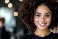 Young African American woman with natural curly hair and bright smile against blurred bokeh background wearing black top, radiating confidence and joy in professional setting.