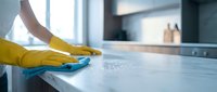 Person in yellow rubber gloves cleaning white kitchen counter with blue cloth, maintaining hygiene and cleanliness in modern home interior.