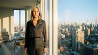 Professional Caucasian businesswoman in dark suit standing by floor-to-ceiling windows overlooking urban skyline during golden hour in modern office.