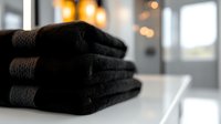 Stack of folded black cotton towels on white surface against blurred modern bathroom interior with soft bokeh lights, creating luxurious spa atmosphere.