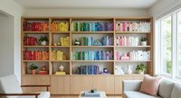 Modern home library with color-coordinated books arranged by rainbow spectrum on built-in wooden shelves, featuring plants and decorative objects in bright interior.