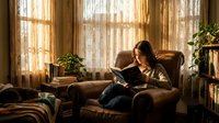 Young woman reading book in cozy armchair by window with warm sunlight streaming through curtains creating peaceful indoor atmosphere for relaxation.