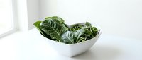 Fresh green leafy vegetables in white bowl on white surface, suitable for healthy eating, nutrition, diet, and food styling projects.