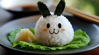 Cute rabbit-shaped rice ball with nori ears and carrot nose served on fresh lettuce leaf. Creative Asian food styling on dark plate with blurred background.