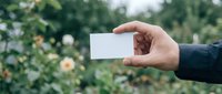 Hand holding blank white business card outdoors with green foliage background for corporate branding and professional networking mockup template design.