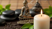 Zen spa composition with burning candle, Buddha statue, hot stones and green leaves on wooden background creating peaceful meditation atmosphere.