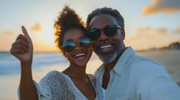 Happy African American couple taking selfie at sunset beach, wearing sunglasses and casual attire, showing genuine smiles and positive emotions during romantic vacation moment.