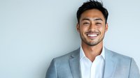 Young Asian businessman with confident smile wearing grey suit and white shirt against light background, professional corporate portrait.