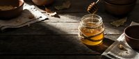 Warm honey dripping from wooden dipper into glass jar on rustic wooden table with autumn leaves and natural lighting for cozy kitchen scenes.