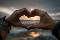 Hands forming heart shape against sunset sky over water creating romantic silhouette for love and relationship concepts.