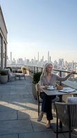 Mature blonde businesswoman enjoying coffee on luxury rooftop terrace with city skyline view during golden hour morning routine.