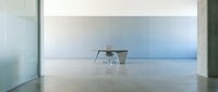 Minimalist interior space with single chair and desk in empty concrete room with large windows overlooking calm water horizon, conveying solitude and modern workspace design.