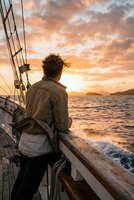 Young man sailing on yacht deck at golden sunset watching ocean waves and dramatic sky during maritime adventure journey.