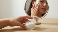 Young adult female, Caucasian ethnicity, applying moisturizing cream to face while looking in tabletop mirror. Skincare routine, cosmetics, and beauty product application for personal care and wellness purposes.