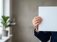 Businessman in dark blue suit holding blank white card covering face, indoor office setting with potted plant and window light, corporate communication concept.