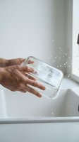 Hands washing a clear glass food container under running water at a white kitchen sink, with splashing droplets, eco-friendly reusable lifestyle concept.