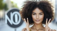 Young African American woman with natural curly afro hair showing peace signs and smiling outdoors, expressing positivity and confidence against urban background with no sign.