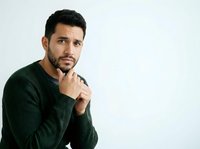 Middle-aged man with dark hair and beard wearing forest green ribbed sweater, hand near chin in thoughtful pose against white background, suitable for business and lifestyle content.