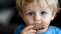 Young caucasian toddler with bright blue eyes and blonde hair wearing blue shirt takes big bite of chocolate cookie while looking at camera with innocent expression.