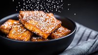 Crispy caramelized salmon fillets with sesame seeds served in black ceramic bowl on dark background. Powdered seasoning falling creates dynamic effect.
