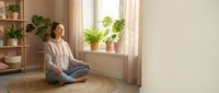 Young woman practicing mindfulness meditation sitting cross-legged on round jute rug in bright living room with houseplants and natural light streaming through window.