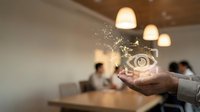 Young male professional holding glowing eye icon with digital particles in modern office workspace with colleagues and pendant lighting for vision, insight, and business technology concepts.