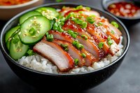 Sliced char siu pork belly served on steamed rice with fresh cucumber and green onions in black bowl, topped with sesame seeds and sweet red sauce.