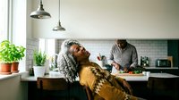 African American woman with curly gray hair relaxing at kitchen island while partner cooks in modern home with subway tile backsplash and pendant lights.