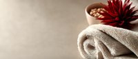 Rolled white spa towel with red tropical flower and bowl of aromatherapy stones on beige background, wellness and relaxation concept. Panoramic composition.