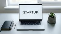 Laptop on a clean white desk displaying the word Startup, with a notebook, pens, and a small plant, symbolizing entrepreneurship and innovation.