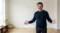 Middle-aged man with dark hair and facial hair, Mediterranean ethnicity, wearing navy sweatshirt and dark pants, standing in bright minimalist interior with arms outstretched in welcoming gesture, suitable for business, lifestyle and motivational content.