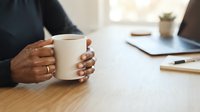 Hands holding warm mug at wooden desk with laptop and notebook, suitable for business, lifestyle, and workplace concepts.