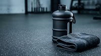 Black water bottle and gray towel on gym floor with blurred fitness equipment in background, suitable for health and wellness marketing.