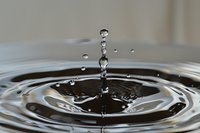 Water drop impact creating concentric ripples and splash crown formation with suspended droplets in mid-air against dark liquid surface. Macro photography captures fluid dynamics.
