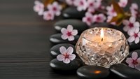 Zen spa setting with lit candle in crystal holder, cherry blossoms and smooth black stones on dark wooden surface for relaxation and wellness treatments.