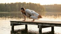Young athletic man practicing yoga crow pose on wooden dock at sunset lake. Fitness meditation balance exercise outdoors in nature during golden hour.