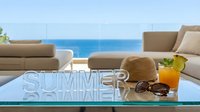Luxury terrace with summer lettering, straw hat, sunglasses, and tropical cocktail on glass table overlooking a serene blue ocean view.