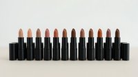 Professional makeup collection featuring diverse nude and brown lipstick shades in black tubes arranged in gradient from light to dark tones.