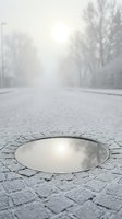 Snowy street with bare trees and fog in background, ground-level view of open manhole cover reflecting overcast winter sky on paved road.