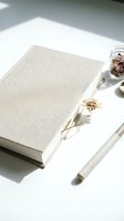 Closed beige linen book with dried flowers and glass jar on white surface, flat lay composition for publishing, stationery, and lifestyle design projects.