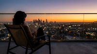 Woman sitting on rooftop chair overlooking illuminated cityscape at sunset, urban skyline view from high-rise balcony during golden hour.