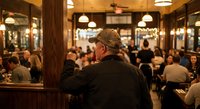 Mature Caucasian man in cap observing busy restaurant interior with warm lighting and dining patrons enjoying evening meal service.